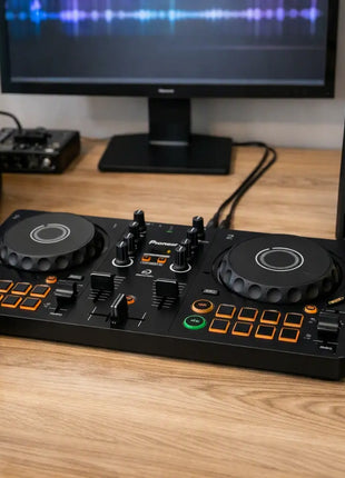 A black Pioneer DJ controller for DJs sits on a wooden desk, flanked by two matching Pioneer DJ studio speakers and a pair of black over-ear headphones. A computer monitor is visible in the background.